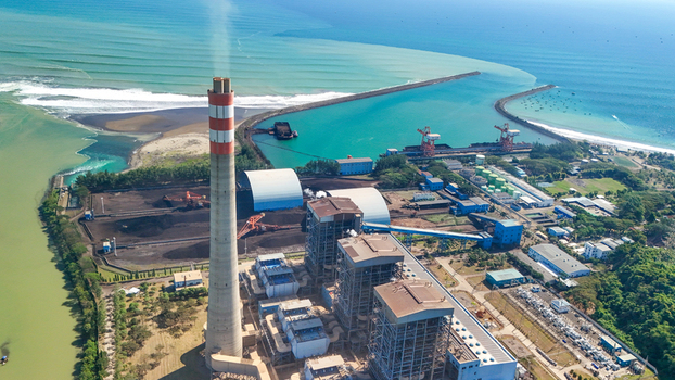 Aerial view of a large coal fired power plant with a tall chimney emitting smoke situated on the coast next to the turquoise sea and a breakwater in Indonesia.