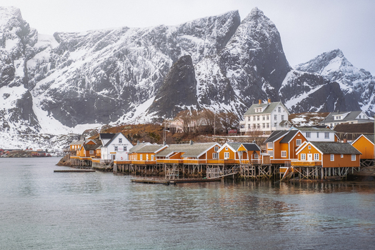 View of golden cabins and white houses nestle by the water s edge contrasting with snow capped mountains under a muted sky Hamn&oslash;y Nordland Norway.