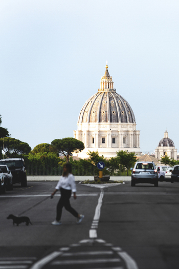 View of St. Peter s Basilica dome rising above the city with a woman walking a dog on a street in Rome Lazio Italy. View of St. Peter s Basilica dome rising above the city with a woman walking a dog on a street in Rome Lazio Italy.