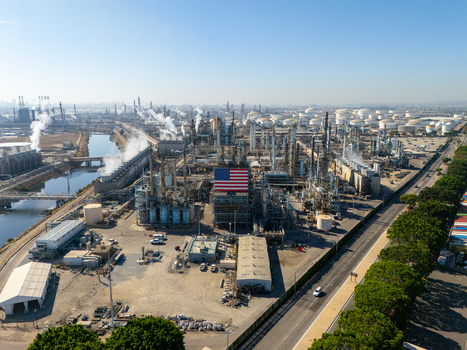 Aerial view of an industrial oil refinery in Carson featuring a large American flag on a central structure storage tanks and steam plumes. Carson California United States.