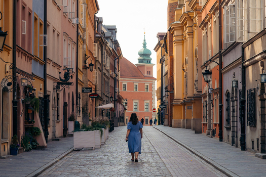 View of Warsaw Old Town with a woman walking along the cobblestone street lined with colorful historic buildings and a church tower in the background in Warsaw Masovian Voivodeship Poland. View of Warsaw Old Town with a woman walking along the cobblestone street lined with colorful historic buildings and a church tower in the background in Warsaw Masovian Voivodeship Poland.