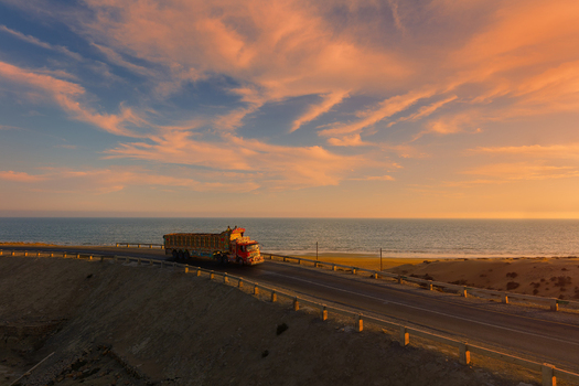 View of a decorated truck driving along the Makran Coastal Highway during a vibrant sunset with the sea and desert landscape in Balochistan Pakistan.