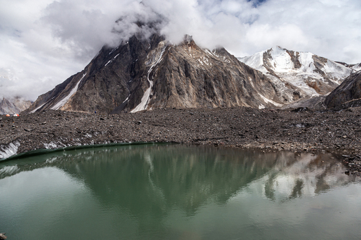 View of the Baltoro Glacier with a glacial lake in the foreground surrounded by rocky moraine and snow capped mountain peaks under a cloudy sky in Baltoro Gilgit Baltistan Pakistan.