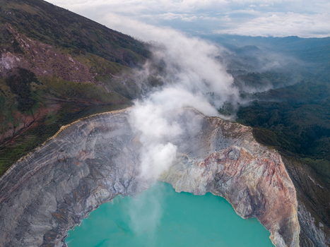 Aerial view of Ijen crater featuring its iconic turquoise acidic lake rugged volcanic walls and rising steam plumes set against the mountainous landscape in Ijen East Java Indonesia.