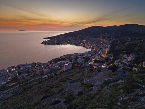 Aerial view of the coastal city of Saranda illuminated by city lights at sunset with the Ionian Sea and surrounding hills in Saranda Vlore County Albania. Aerial view of the coastal city of Saranda illuminated by city lights at sunset with the Ionian Sea and surrounding hills in Saranda Vlore County Albania.