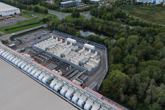 Aerial view of a data center with cooling units and industrial infrastructure surrounded by trees and a pond in the United Kingdom.