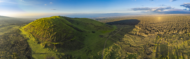 Aerial view of the Puu Waawaa cinder cone featuring lush green slopes and surrounding volcanic lava fields under a clear sky in Hawaii County Hawaii United States. Aerial view of the Puu Waawaa cinder cone featuring lush green slopes and surrounding volcanic lava fields under a clear sky in Hawaii County Hawaii United States.