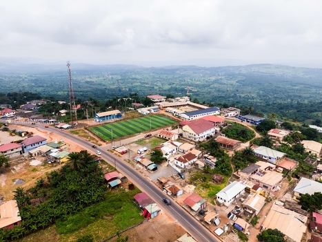 Aerial view of a small town featuring a green football field a paved road scattered residential buildings and a communication tower set against a hilly landscape in Ghana. Aerial view of a small town featuring a green football field a paved road scattered residential buildings and a communication tower set against a hilly landscape in Ghana.