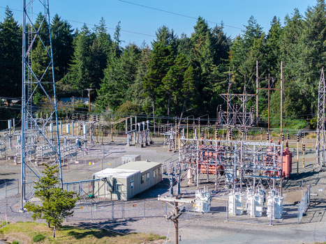 Aerial view of an electrical substation featuring high voltage transformers metal transmission towers and a small utility building surrounded by a dense forest Bandon Oregon United States. Aerial view of an electrical substation featuring high voltage transformers metal transmission towers and a small utility building surrounded by a dense forest Bandon Oregon United States.