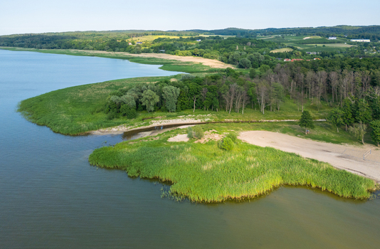 Aerial view of the Vistula Lagoon coastline featuring lush green reeds a sandy beach and dense forest bordering the water in Tolkmicko Warmian Masurian Voivodeship Poland. Aerial view of the Vistula Lagoon coastline featuring lush green reeds a sandy beach and dense forest bordering the water in Tolkmicko Warmian Masurian Voivodeship Poland.