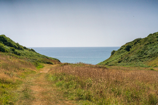 View of the coastal path leading to Lansallos beach framed by grassy hills and green cliffs overlooking the calm blue sea in Lansallos England United Kingdom. View of the coastal path leading to Lansallos beach framed by grassy hills and green cliffs overlooking the calm blue sea in Lansallos England United Kingdom.