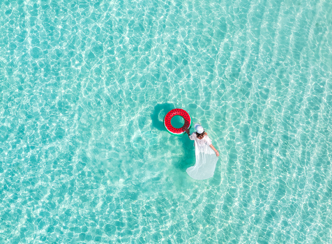 Aerial view of a woman in a white dress standing in crystal clear turquoise water with a watermelon inflatable ring Vashafaru Haa Alif Atoll Maldives. Aerial view of a woman in a white dress standing in crystal clear turquoise water with a watermelon inflatable ring Vashafaru Haa Alif Atoll Maldives.