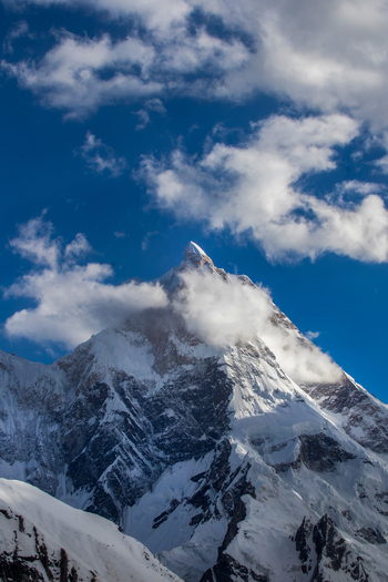 View of the snow covered peak of Masherbrum mountain partially hidden by white clouds under a blue sky in Skardu Gilgit Pakistan. View of the snow covered peak of Masherbrum mountain partially hidden by white clouds under a blue sky in Skardu Gilgit Pakistan.