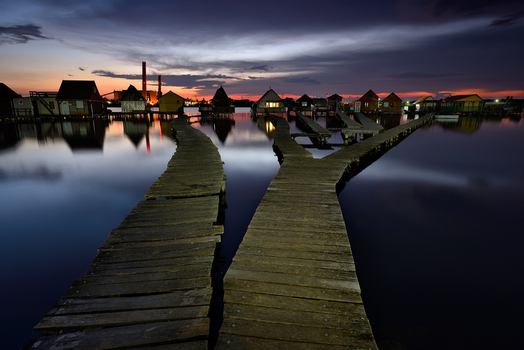 View of wooden stilt houses on a calm lake with wooden walkways leading to them under a dramatic sunset sky in Hungary.