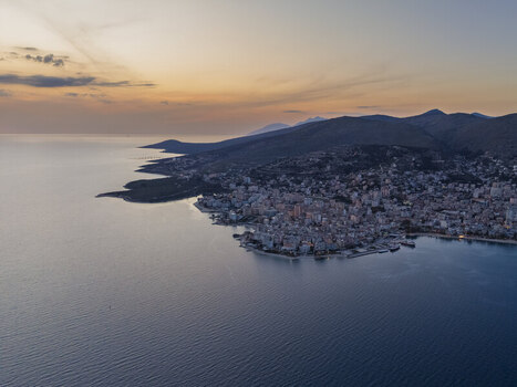 Aerial view of the coastal city of Saranda at sunset featuring urban buildings along the shoreline calm sea and rolling mountains in Saranda Vlore County Albania. Aerial view of the coastal city of Saranda at sunset featuring urban buildings along the shoreline calm sea and rolling mountains in Saranda Vlore County Albania.