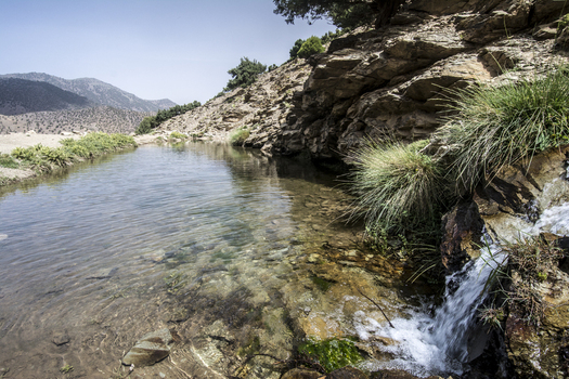 View of a clear mountain stream and small waterfall flowing through rocky terrain with sparse green vegetation in the High Atlas mountains Tizi Oussem Marrakech Safi Morocco. View of a clear mountain stream and small waterfall flowing through rocky terrain with sparse green vegetation in the High Atlas mountains Tizi Oussem Marrakech Safi Morocco.
