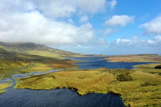 Aerial view of The Storr a dramatic rocky landscape with rolling green hills dark blue lochs and scattered clouds under a bright sky in Portree Scotland United Kingdom.