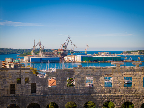 Aerial view of the ancient Roman amphitheater s weathered stone facade framing the bustling port with towering cranes against the azure sea Pula Istria County Croatia.