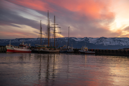 View of serene harbor reflecting a vibrant sunset sky with boats gently resting against the backdrop of snow capped mountains Húsavík Norðurþing Iceland. View of serene harbor reflecting a vibrant sunset sky with boats gently resting against the backdrop of snow capped mountains Húsavík Norðurþing Iceland.
