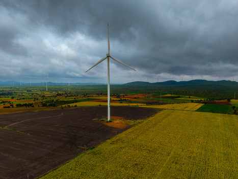 Aerial view of a wind turbine standing in agricultural fields with yellow crops and dark soil under a cloudy sky in Hampi Karnataka India.
