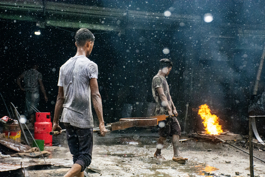 Keraniganj Bangladesh 28 August 2022 View of workers carrying a heavy metal component in a shipyard with a glowing fire and industrial equipment.