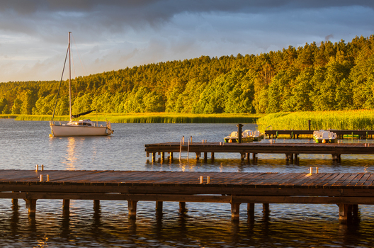 View of a sailboat moored near wooden piers on a calm lake surrounded by lush forest during golden hour in Male Swornegacie Pomeranian Voivodeship Poland.