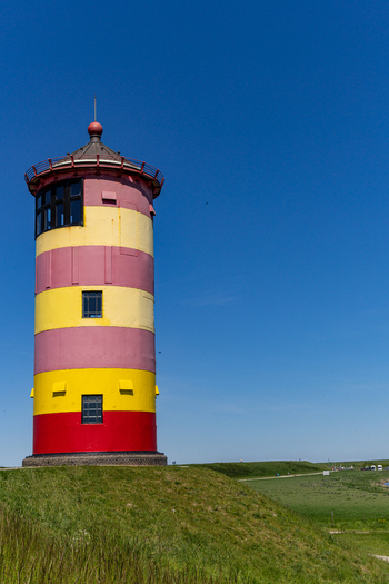 View of the iconic Pilsum lighthouse with its distinctive yellow and red stripes standing on a grassy dike under a clear blue sky in Krummhorn Niedersachsen Germany. View of the iconic Pilsum lighthouse with its distinctive yellow and red stripes standing on a grassy dike under a clear blue sky in Krummhorn Niedersachsen Germany.