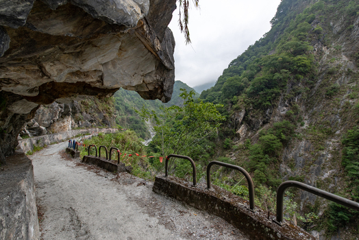 View of a narrow unpaved cliffside road with metal guardrails winding through the steep lush forested mountains of Taroko National Park in Xiulin Township Hualien County Taiwan.