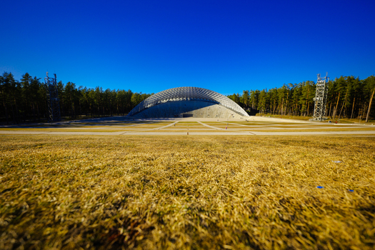 Aerial view of the Mezaparks Open Air Stage featuring its distinctive arched roof structure surrounded by a pine forest under a clear blue sky in Riga Latvia. Aerial view of the Mezaparks Open Air Stage featuring its distinctive arched roof structure surrounded by a pine forest under a clear blue sky in Riga Latvia.