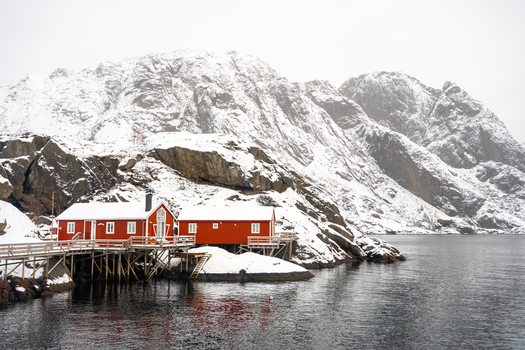 View of traditional red wooden cabins on stilts over the water against a backdrop of snow covered mountains in Leknes Lofoten Norway.