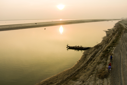 Aerial view of a traditional wooden boat on the calm Padma River at sunset with people walking along the sandy riverbank in Rajshahi Rajshahi Division Bangladesh.