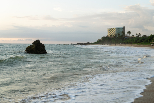 View of the sandy coastline with waves crashing near a large rock formation and a modern hotel building in the distance Galle Southern Province Sri Lanka.