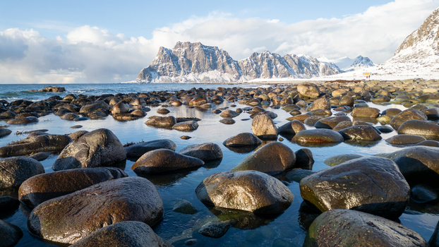 View of a rocky beach with smooth boulders in the foreground and snow capped mountains under a clear sky in Leknes Lofoten Norway.