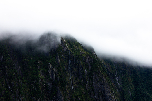 View of the misty towering peaks rise majestically shrouded in ethereal clouds their lush green slopes contrasting with the stark grey rock Milford Sound Southland Region New Zealand.