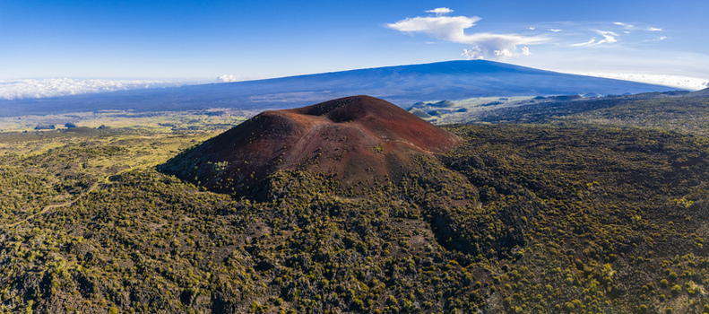 Aerial view of the Mauna Kea volcano and a reddish cinder cone surrounded by rugged lava fields and sparse vegetation under a clear blue sky in Hawaii County Hawaii United States.