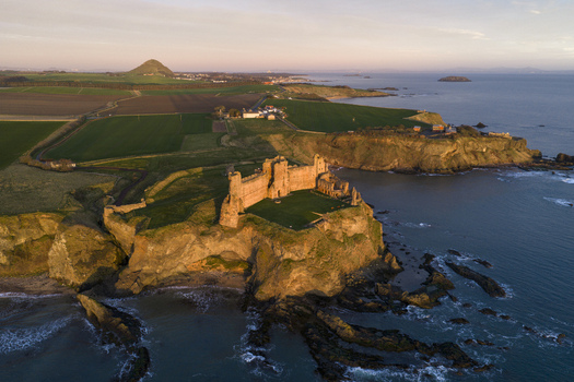 Aerial view of Tantallon Castle on a rocky cliff overlooking the North Sea with green fields and North Berwick Law in the distance during golden hour North Berwick Scotland United Kingdom.