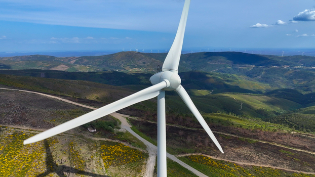 Aerial view of a wind turbine standing on a green hillside with yellow wildflowers and a dirt road under a clear blue sky in Lisbon Lisbon Portugal. Aerial view of a wind turbine standing on a green hillside with yellow wildflowers and a dirt road under a clear blue sky in Lisbon Lisbon Portugal.