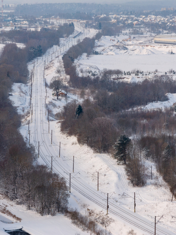 Aerial view of snow covered railway tracks winding through a winter forest landscape with bare trees and distant industrial buildings Lithuania. Aerial view of snow covered railway tracks winding through a winter forest landscape with bare trees and distant industrial buildings Lithuania.