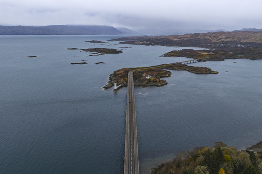 Aerial view of the Skye Bridge spanning across the sea with a small lighthouse and rocky islands under an overcast sky Isle of Skye Scotland United Kingdom.