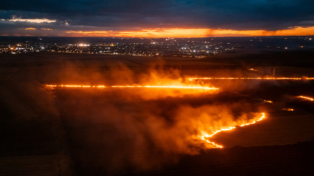 Aerial view of a field fire burning at night with bright orange flames and smoke set against a dark landscape with distant city lights in Sremska Mitrovica Vojvodina Serbia.
