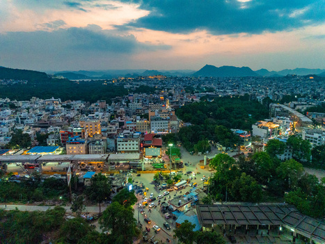 Aerial view of the urban landscape at dusk featuring dense buildings busy roads with traffic lush green trees and distant mountains Udaipur Rajasthan India. Aerial view of the urban landscape at dusk featuring dense buildings busy roads with traffic lush green trees and distant mountains Udaipur Rajasthan India.