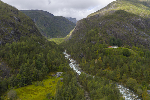 Aerial view of a winding river flowing through a lush green valley surrounded by steep forested mountains and scattered rural houses in Fjaera Vestland Norway. Aerial view of a winding river flowing through a lush green valley surrounded by steep forested mountains and scattered rural houses in Fjaera Vestland Norway.