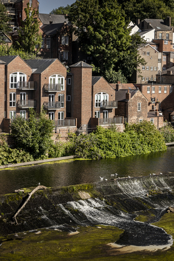 View of the River Wear weir with historic brick buildings and lush greenery along the riverbank in Durham England United Kingdom. View of the River Wear weir with historic brick buildings and lush greenery along the riverbank in Durham England United Kingdom.