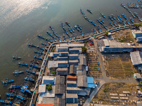 Aerial view of a coastal fishing village with numerous small wooden boats moored along the shore featuring simple houses and drying racks in Cilacap Regency Central Java Indonesia. Aerial view of a coastal fishing village with numerous small wooden boats moored along the shore featuring simple houses and drying racks in Cilacap Regency Central Java Indonesia.