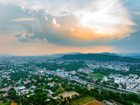 Aerial view of the cityscape of Udaipur with a railway station lush green trees and distant hills under a dramatic sunset sky in Udaipur Rajasthan India. Aerial view of the cityscape of Udaipur with a railway station lush green trees and distant hills under a dramatic sunset sky in Udaipur Rajasthan India.