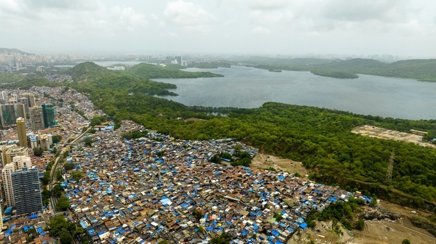 Aerial view of Dharavi Slums with dense residential rooftops adjacent lush green hills and the expansive Powai Lake in the background Mumbai Maharashtra India. Aerial view of Dharavi Slums with dense residential rooftops adjacent lush green hills and the expansive Powai Lake in the background Mumbai Maharashtra India.