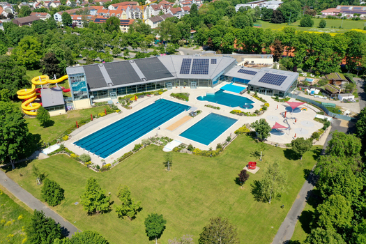 Aerial view of Konigsbad Forchheim featuring outdoor swimming pools yellow water slides and solar panels on the roof surrounded by lush green trees in Forchheim Bayern Germany. Aerial view of Konigsbad Forchheim featuring outdoor swimming pools yellow water slides and solar panels on the roof surrounded by lush green trees in Forchheim Bayern Germany.