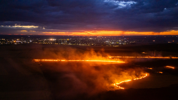 Aerial view of a field fire burning at night with glowing flames and smoke set against the backdrop of the illuminated city of Sremska Mitrovica Vojvodina Serbia.