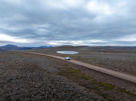 Aerial view of a solitary car navigates a gravel path through the stark rocky landscape punctuated by a small lake under a vast overcast sky Westfjords Iceland. Aerial view of a solitary car navigates a gravel path through the stark rocky landscape punctuated by a small lake under a vast overcast sky Westfjords Iceland.