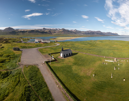 Aerial view of the iconic black Budakirkja church situated in a green field with a cemetery gravel path and coastal landscape in the background in Budir Snaefellsbaer Iceland. Aerial view of the iconic black Budakirkja church situated in a green field with a cemetery gravel path and coastal landscape in the background in Budir Snaefellsbaer Iceland.